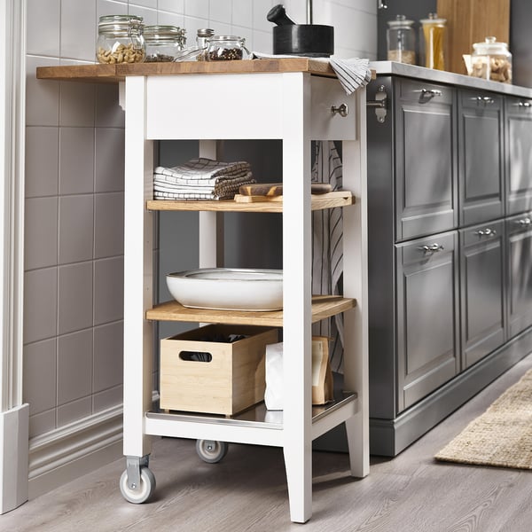 White STENSTORP kitchen trolley with wooden shelves. Stores jars, towels, and bowls. Beside cabinets and tiled wall.