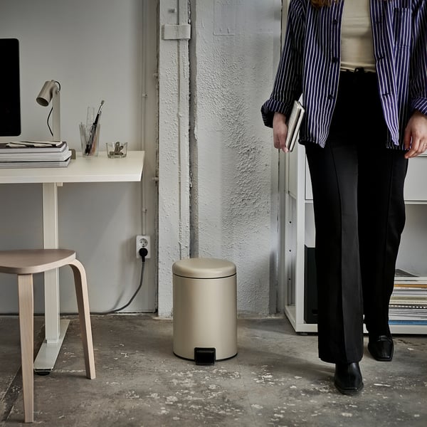 Person holding books near simple home office setup with computer, small rubbish can, and stool.