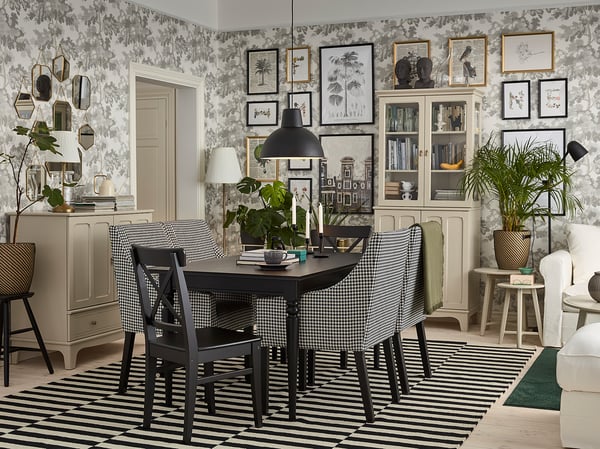 Modern dining room a black dining table with black chairs, striped rug, grey cabinets, plant, & artistic wall decor with floral wallpaper.