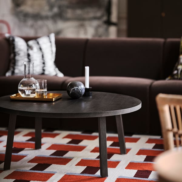 Dark brown STOCKHOLM 2025 coffee table with oak top on colourful rug, holding decanter, glasses, and a candle. Larger than nearby wooden chair.