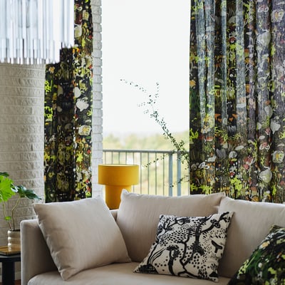 Bright living room with large, patterned stockholm 25 curtains framing a window, a beige couch with decorative pillows, and a yellow lamp beside it.