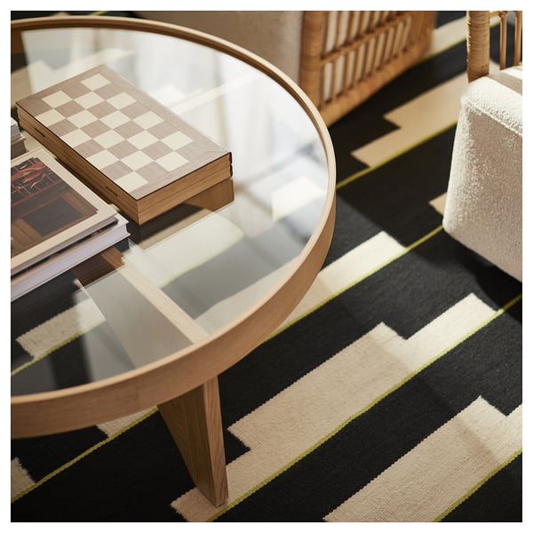 Round glass coffee table with wooden base, books, and chess board on striped rug.