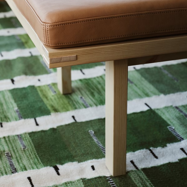 A beige leather stool with light wood legs sits on a green handwoven rug with a birch tree pattern, emphasising comfort and style.