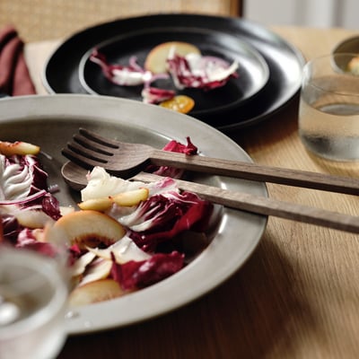 Beige serving bowl with rustic salad and wooden tongs, on wooden table.