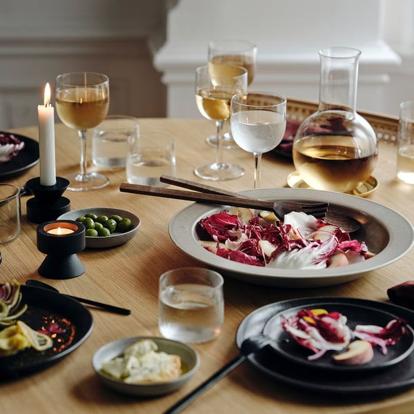 Wooden table set with beige stoneware dishes, colourful salad, candles, wine glasses, and carafe with white wine. Centrepiece with wooden utensils.