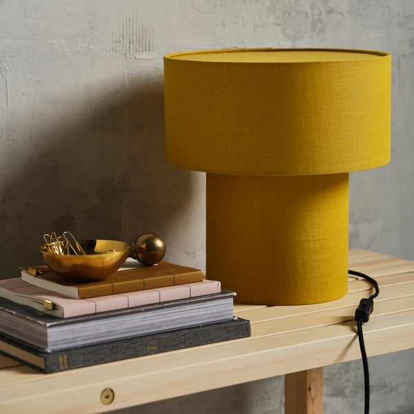 Bright yellow STOCKHOLM 2025 lamp on wood shelf, surrounded by books, offering warm light.
