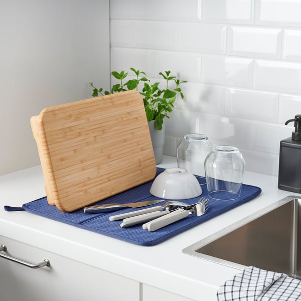 Kitchen scene with cutting board, knives, bowl, vases, and herbs on blue mat near sink.