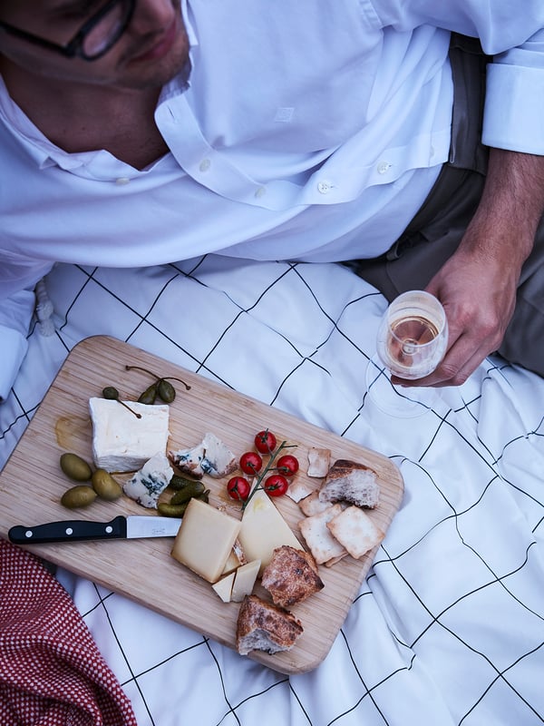 Person in white shirt holding wine glass near wooden board with cheese, olives, cherry tomatoes, and knife on blue chequered cloth.