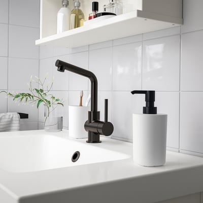 Modern bathroom sink with STORAVAN black tap and white accessories. Pump dispenser, toothbrush holder, and tray on clean white countertop.