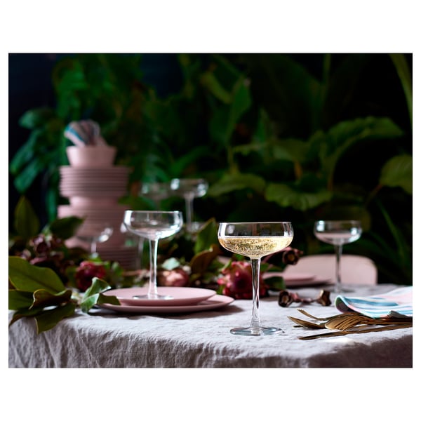 Beautiful table setting with clear, wide-mouthed wine glasses on a linen tablecloth, surrounded by pink plates, gold cutlery, and lush green plants.