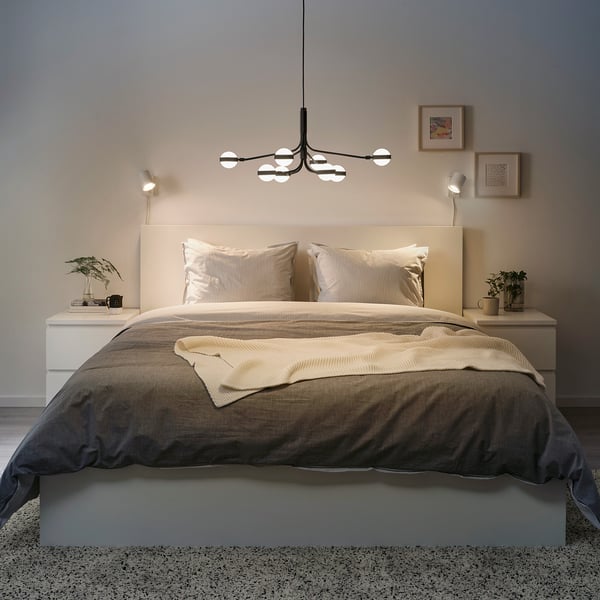 Modern bedroom with STORSLINGA chandelier above bed, soft lighting, neutral linens, plants, minimalist art, cozy ambiance.