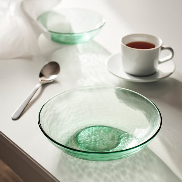 Glass plate named STRANDKRABBA in green, hammered design, round & large, sits near a white coffee cup and spoon on a light table.