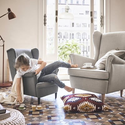 Cosy living room with a child on the STRANDMON kids armchair. Light grey, comfy seat, large floor cushion, cups, rug.