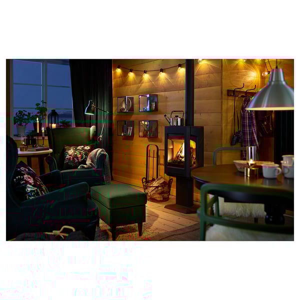 Cosy living room with green STRANDMON chairs, wooden walls, lit fireplace, and string lights.