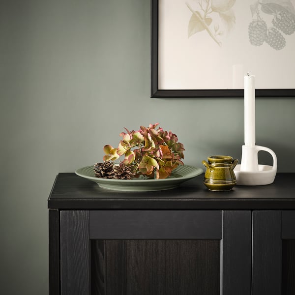 Green ceramic plate with autumn leaves and pinecones, jug, and candle holder against a grey wall.