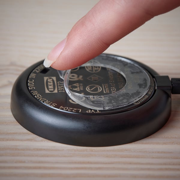 Hand pressing black STRIMSÄV spotlight on wood, powered by gel pad.