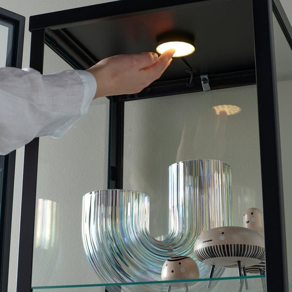 Person pressing a spotlight mounted inside a glass cabinet. The spotlight illuminates decorative glass vases.