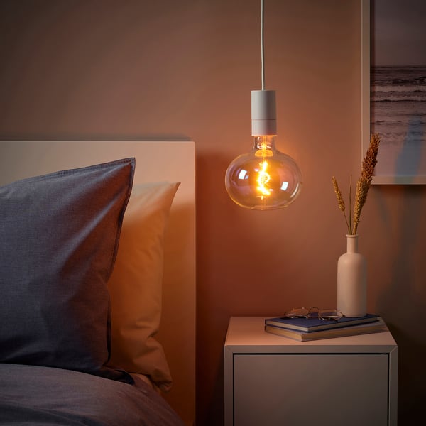 SUNNEBY lamp above bedside table in cosy room. Peach walls, mixed pillows, neat cord, books, vase with dried grasses. Sleek, minimalist design.