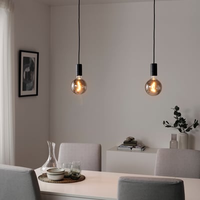 Two smoke glass pendants over table, warm ambiance. Woven chairs, wall art, vase with dried branches.