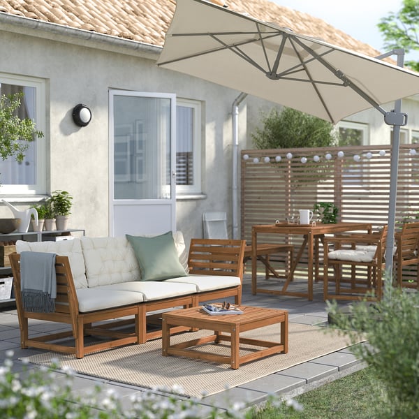 Backyard patio with wooden furniture, white cushions, large beige umbrella, and string lights.