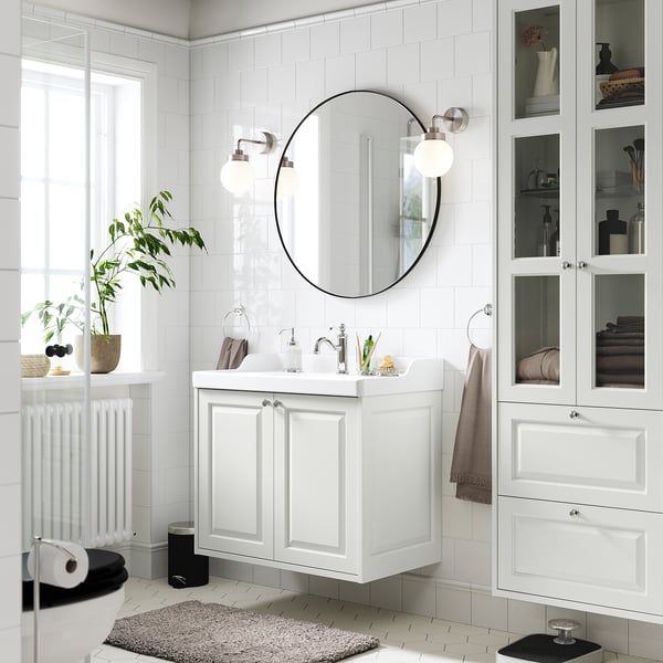 Modern white bathroom with TÄNNFORSEN vanity, large round mirror, glass-front cabinets, and radiator. Plants and towels enhance the minimalist design.
