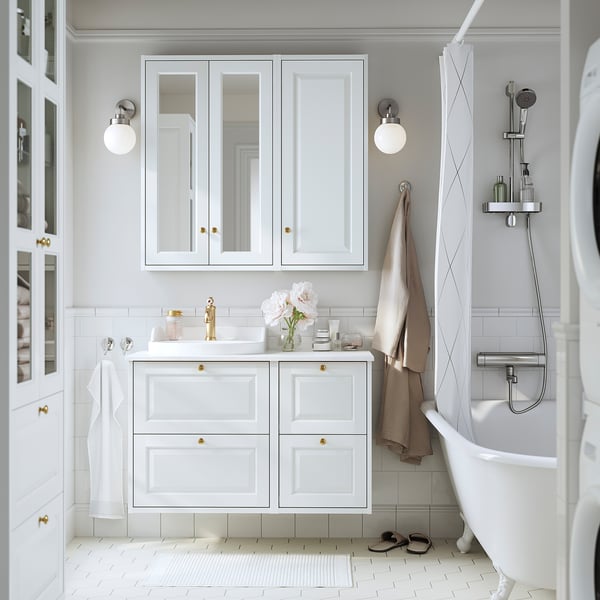 Modern white bathroom with TÄNNFORSEN cabinet, gold accents, glass shelf, and flowers. Shallow cabinet above sink, fitting well between wall lights.