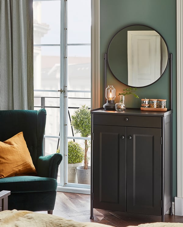 Cosy room with green walls, a black TÄRNABY lamp on a wooden cabinet, a round mirror, and a teal chair by a window.