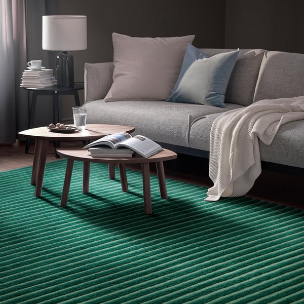Cosy living room with grey sofa, lavender/blue pillows, side tables with water & magazine, dark grey walls, green striped rug.