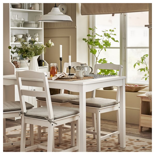 White dining set with cushioned chairs, includes table, teapot, and candles. Modern farmhouse style with open shelving and rug.