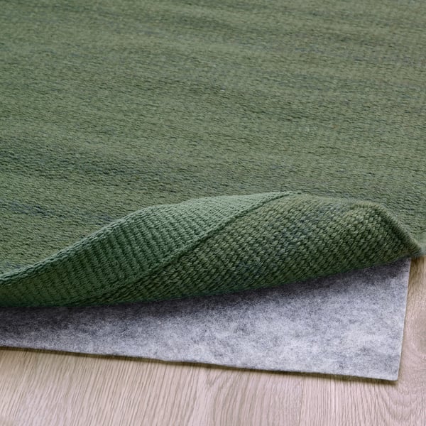 Green flatwoven rug on wooden floor, edge folded showing underside with anti-slip pad.