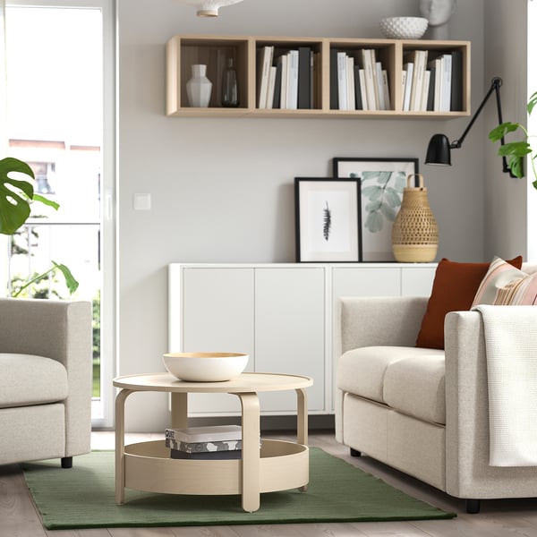 A cozy living room with a TIDTABELL light green rug, neutral furniture, cotton wool, coffee table, chairs, bookshelf, plants, and lamp.