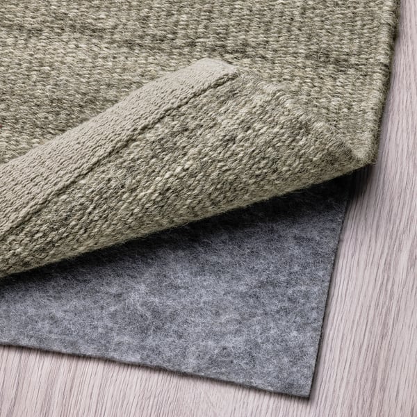 A folded grey wool rug is displayed, showcasing its underside with an anti-slip felt pad attached, ensuring stability and comfort.