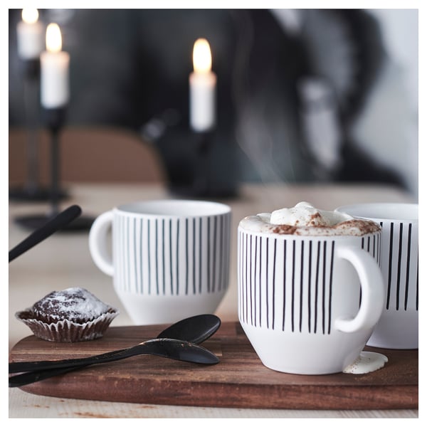 Two white mugs with black stripes and whipped cream drinks, one spoon, one fork, and one knife on a wooden table with lit candles in the background.