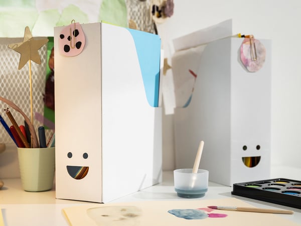 Two white, smiling TJABBA magazine files made from 100% recycled corrugated board on a table with art supplies.