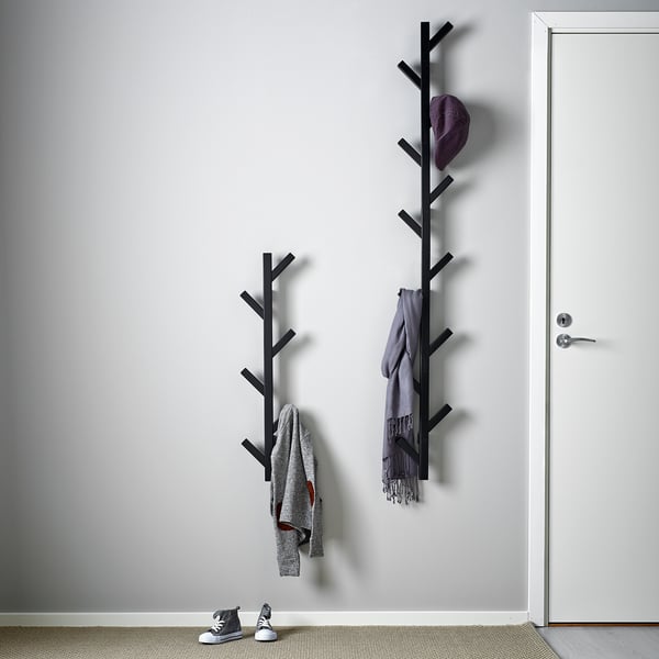 Two wooden coat racks mounted on a white wall hold a grey jacket, blue scarf, and purple hat. A pair of black and white trainers sits below.