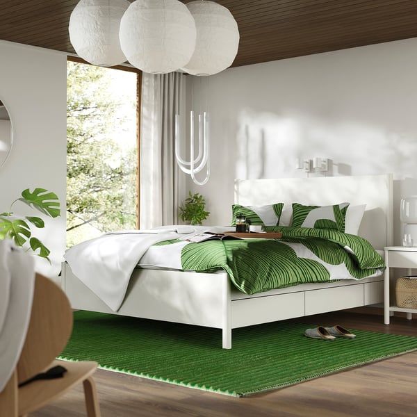 Modern white bed with green rug & pillows, large window, hanging white lamps.