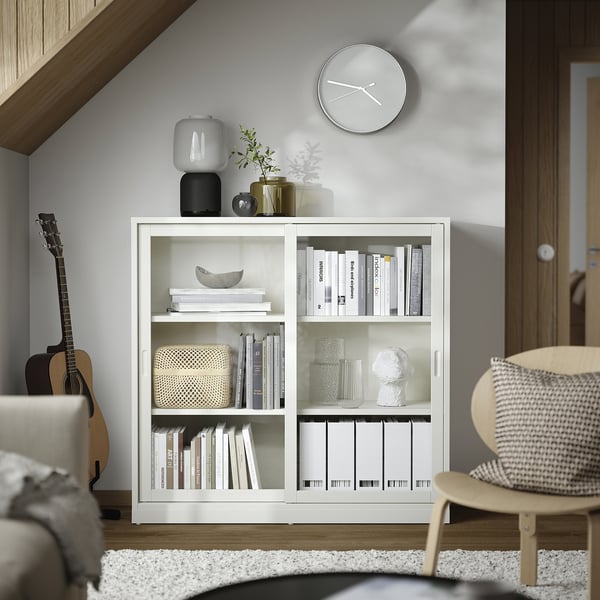 Minimalist living room with white TONSTAD bookshelf, featuring sliding doors, glass vases, stacked decor, and wall clock.