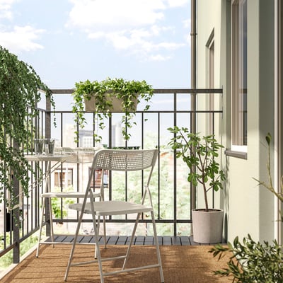 Cosy balcony with TORPARÖ chairs and table on small rug, two green plants, beige wall with window.