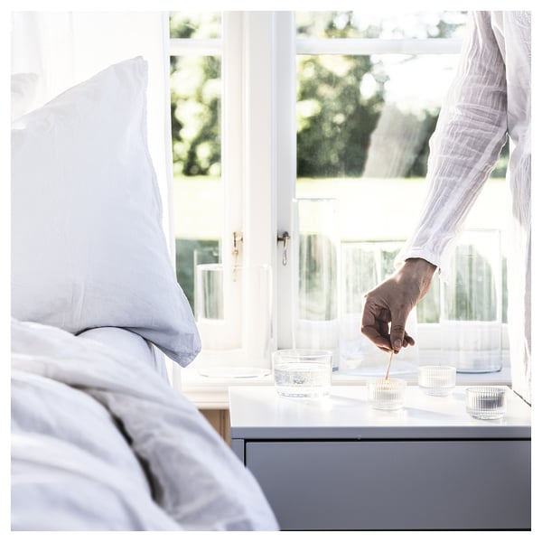 A hand lights a small glass TRÅDCYPRESS candle on a windowsill beside an unmade bed, outside view.