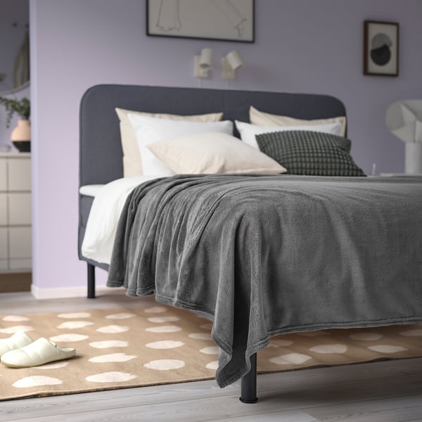 A cosy bed with grey TRATTVIVA bedspread, white pillows, green cushion, purple walls, rug, and slippers.