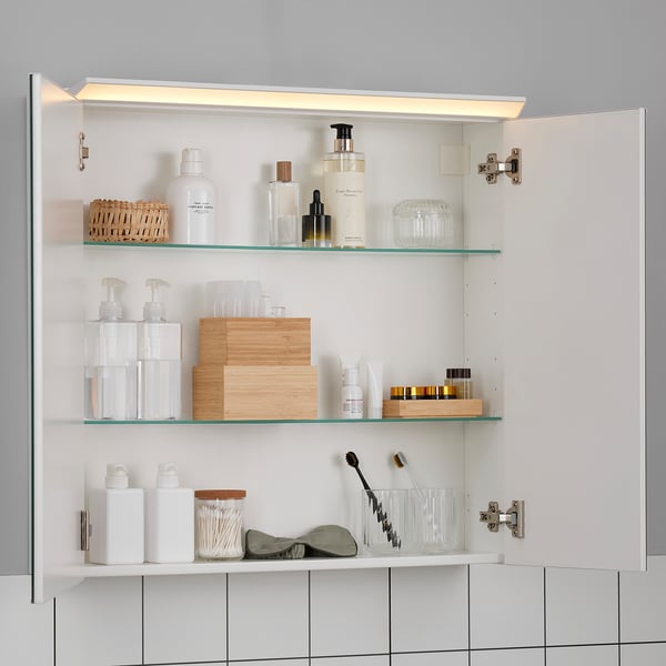 Open bathroom cabinet with light, mirrored doors revealing glass shelves stocked with toiletries, wooden boxes, and personal care items.