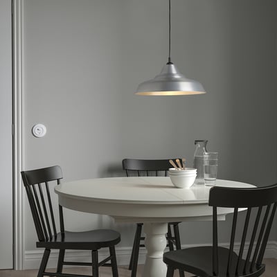 Minimalist dining room with round white table, four black chairs, and TRETTIOEN aluminium lamp. Soft lighting, light grey walls, and cosy vibe.