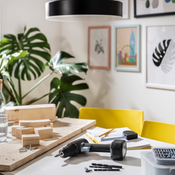 A desk with diy tools: a drill, hammer, screws, wooden pieces, and a plant. Artwork hangs on the wall.