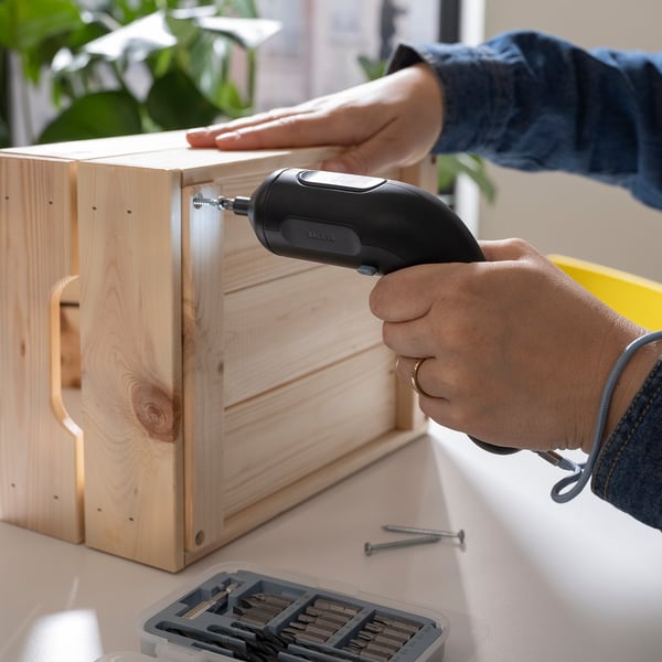 A person uses a cordless screwdriver, the TRIXIG, to assemble wooden furniture. The magnetic bit holder secures the tool in place.