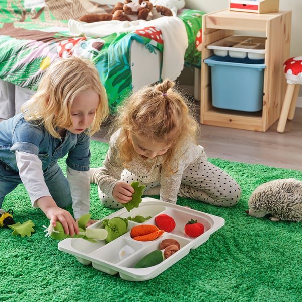 Two young girls play with a TROFAST storage tray filled with colourful plastic vegetables on a green rug. They sort items into compartments.