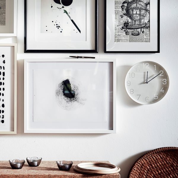 Wall art frames and a white clock. Clocks face displays numbers as dots, black hands show time. Below, small bowls sit on a woven basket and table.