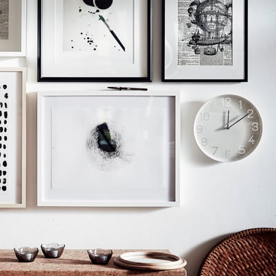 Wall art frames and a white clock. Clocks face displays numbers as dots, black hands show time. Below, small bowls sit on a woven basket and table.
