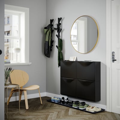 Black modern shoe cabinet, TRONES by IKEA, with a round gold mirror and wooden hanger rack.