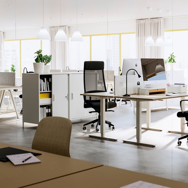 Modern office space with adjustable desks, chairs, and storage units, featuring large windows and bright lighting.