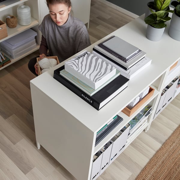 A person sits near a large TROTTEN white double-sided cabinet, taller than the person, with various styled books stacked on top.